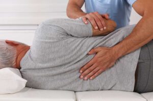 Adult seated in a bright room while a clinician evaluates neck and shoulder movement.
