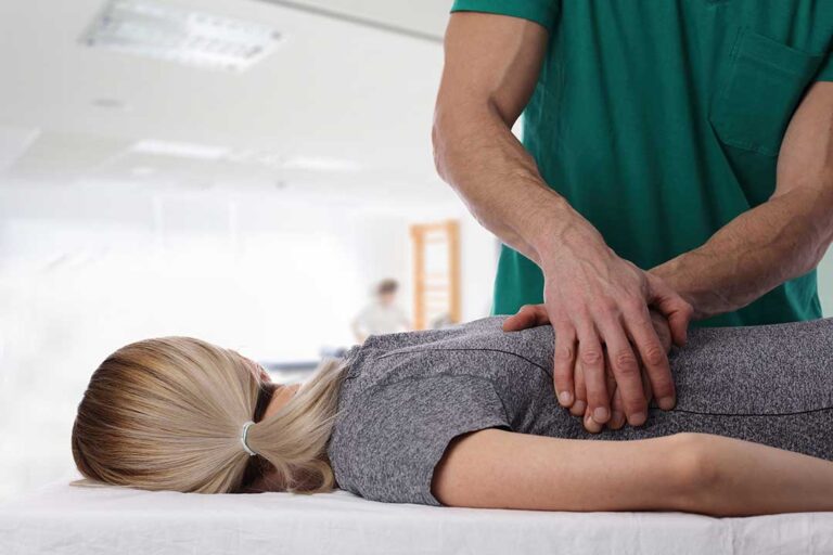 Clinician observing a person’s posture during a gentle scoliosis assessment in a bright room.
