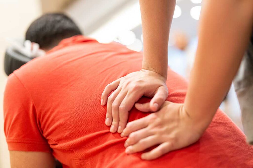 Person gently stretching a sore back beside a treatment table while discussing symptoms with a healthcare provider.